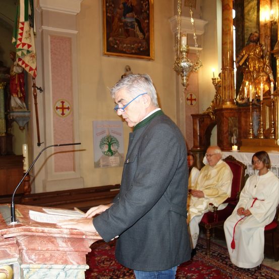 Ein Mann steht an einem Pult und liest ein Buch, mit Brille, in einer Kirche. Hinter ihm sitzen ein Priester und eine Nonne auf Stühlen. Kerzen und ein Kreuz schmücken den Hintergrund.