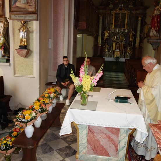 Ein Priester steht und liest aus einem Buch an einem mit einem weißen Tuch bedeckten Tisch, und zwei Männer sitzen auf einer Bank in der Nähe. Auf der linken Seite stehen Vasen mit Blumen.