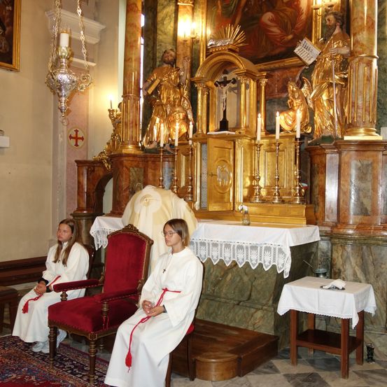 Zwei junge Frauen sitzen in einer Kirche und tragen weiße Gewänder. Sie sitzen vor einem Altar mit goldenen Statuen und brennenden Kerzen. Ein Priester steht hinter ihnen und sieht dem Altar zu. Der Altar hat ein Kreuz und einen Tisch mit einem weißen Tuch.