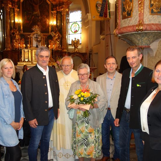 Eine Gruppe von Menschen steht in einer Kirche. Eine ältere Frau mit Blumen in der Mitte und ein Priester auf ihrer rechten Seite. Andere Menschen stehen um sie herum.