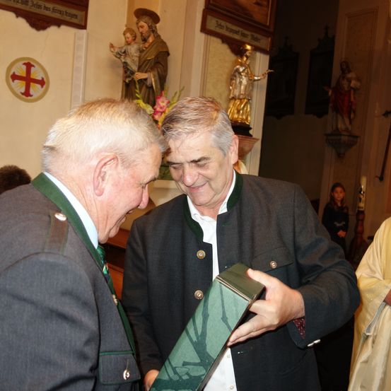 Zwei Männer stehen in einer Kirche, einer hält eine Geschenkbox und schaut den anderen an. Dahinter stehen Statuen und Kerzen.