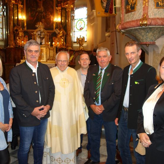 Eine Gruppe von Männern und einer Frau steht in einer Kirche und posiert für ein Foto. Die Männer tragen formelle Kleidung, einer in einem weißen Gewand. Hinter ihnen steht ein Altar mit Kerzen und religiösen Artefakten.
