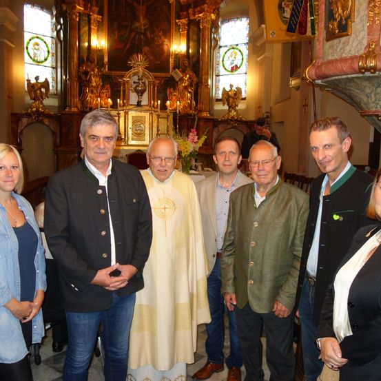Eine Gruppe von Menschen steht in einer Kirche. Ein Priester in einer weißen Robe steht zwischen zwei Männern in Anzügen. Eine Frau in einer Jeansjacke steht links.