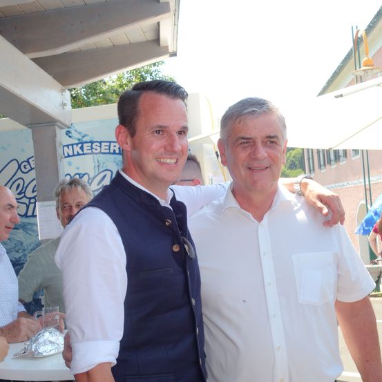 Zwei Männer stehen draußen, einer in einer Weste und der andere in einem weißen Hemd. Sie lächeln und scheinen für ein Foto zu posieren. Hinter ihnen sitzen andere Leute an Tischen.