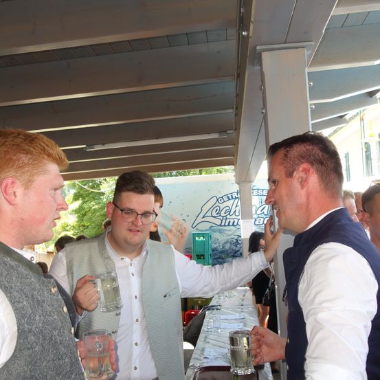 Drei Männer unterhalten sich unter einem Vordach, einer hält ein Glas Wasser. Der Mann auf der rechten Seite scheint eine humorvolle Geste zu machen. Der Hintergrund zeigt einen Tisch mit Gläsern und ein Werbeschild.