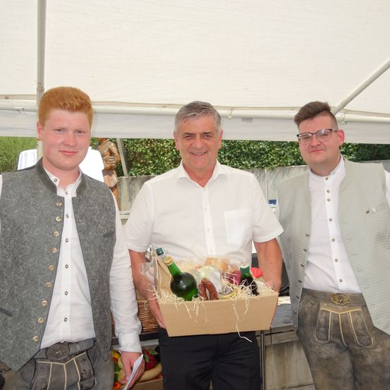 Drei Männer unter einem Zelt, einer hält eine Box mit Essen und Getränken, die anderen lächeln, einer trägt eine Brille.
