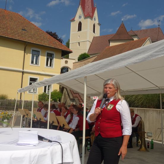 Eine Frau spricht in ein Mikrofon vor einem Tisch unter einem Zelt. Hinter ihr sitzen und spielen eine Gruppe von Menschen Instrumente. Ein Kirchengebäude ist im Hintergrund zu sehen.