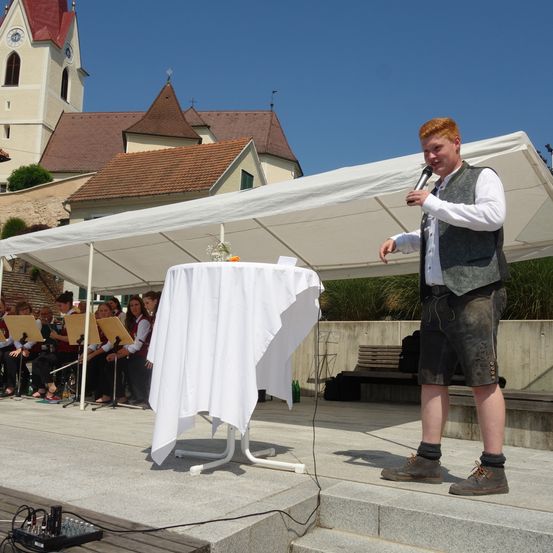 Ein Mann mit roten Haaren steht auf der Bühne und singt mit einem Mikrofon. Hinter ihm steht ein Tisch mit weißer Tischdecke und ein weißes Zelt. Eine Band sitzt vor dem Zelt. Im Hintergrund befinden sich eine Kirche und ein blauer Himmel.