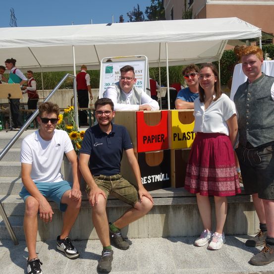 Fünf Menschen sitzen lächelnd auf einer Treppe unter einem Zelt. Dahinter hält ein Mann ein Mikrofon, und zwei Personen stehen in der Nähe eines Tisches. Das Gebiet ist von Pflanzen umgeben.