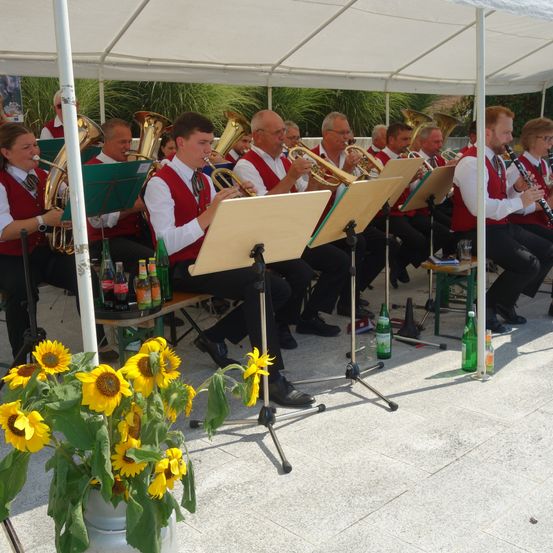 Ein Blasorchester in roten Westen spielt unter einem weißen Zelt mit Sonnenblumen im Vordergrund. Flaschen mit Getränken stehen auf einem Tisch an der Seite.