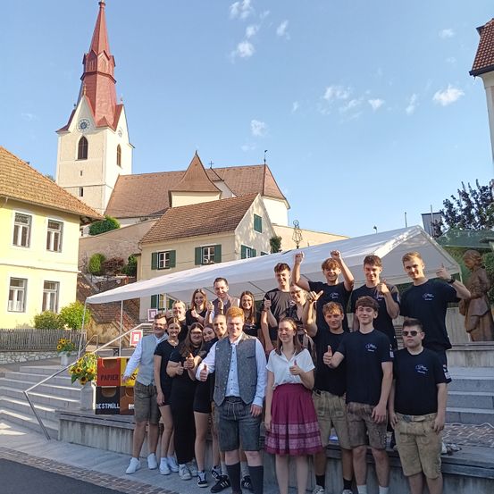 Eine Gruppe von Menschen posiert für ein Foto vor einer Kirche. Sie tragen traditionelle Kleidung und geben Daumen hoch. Die Kirche hat einen Turm mit einer Uhr.
