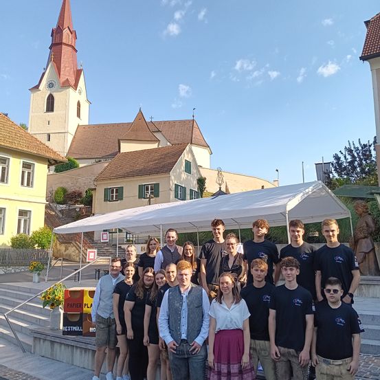 Eine Gruppe von Menschen in gleichen Shirts posiert für ein Foto unter einem weißen Zelt vor einer Kirche. Dahinter sind Stufen, ein Blumentopf und ein Gebäude mit rotem Dach. In der Ferne sind eine Statue, eine Bank und ein Baum zu sehen.