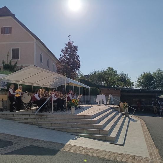 Eine Brassband spielt auf einer Bühne unter einem weißen Zelt. Die Bühne hat Stufen und steht vor einem Gebäude mit einer Ziegelmauer. Im Hintergrund sind Bäume und Menschen zu sehen.