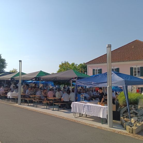 Ein Freisitzbereich mit mehreren Tischen unter Zeltdächern, Menschen versammelt für eine Veranstaltung, mit einem Gebäude im Hintergrund und klarem Himmel.