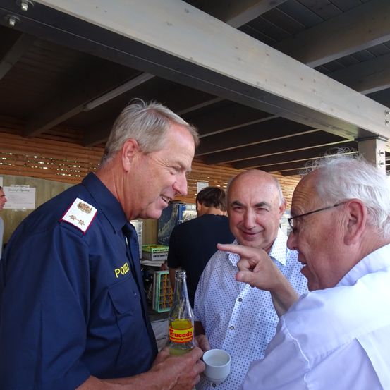 Drei Männer versammeln sich in einem Raum, zwei tragen weiße Hemden und einer ist in einer marineblauen Uniform. Sie führen ein Gespräch. Der Mann in der Marineuniform hält eine Flasche 'Trucade'. Der Mann auf der rechten Seite zeigt auf etwas.