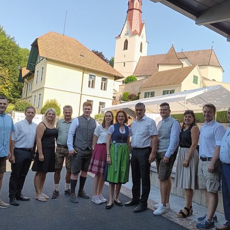 Eine Gruppe von Menschen steht vor einer Kirche mit einem Turm in traditioneller bayerischer Kleidung. Das Gebäude ist gelb mit braunen Dächern, und der Himmel ist klar.
