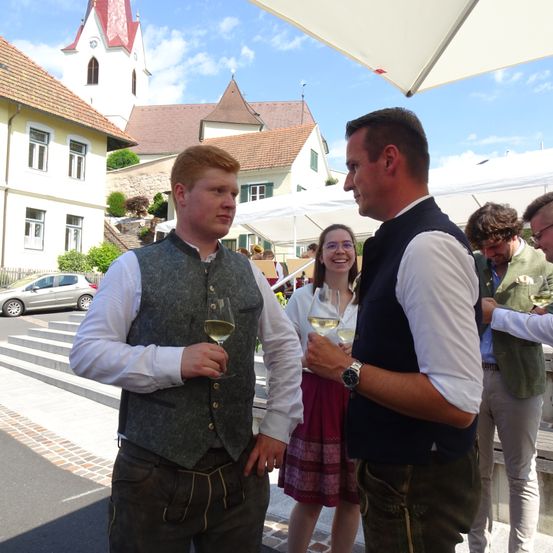 Mehrere Menschen unterhalten sich unter einem Zelt. Ein Mann mit einer Uhr spricht mit einem anderen Mann, der ein Glas Wein in der Hand hält. Eine Frau in einem Rock und mit Brille steht in der Nähe.