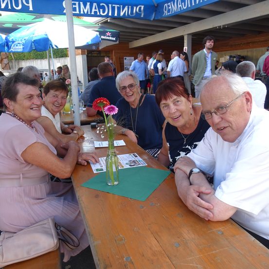 Eine Gruppe von Menschen, hauptsächlich Frauen, sitzt an einem Tisch in einem Café im Freien. Der Tisch hat eine Vase mit Blumen und grünem Papier. Die Menschen lächeln.