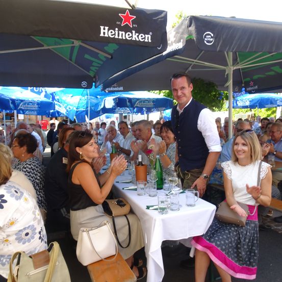 Eine Gruppe von Menschen sitzt unter Regenschirmen, wobei zwei Frauen vor einem Tisch mit Getränken und Gläsern klatschen.