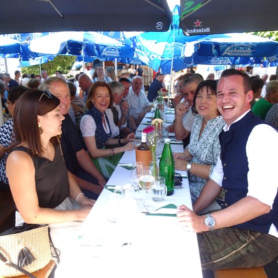 Eine Gruppe von Menschen sitzt an einem langen Tisch im Freien unter blauen Sonnenschirmen. Sie lächeln und lachen. Der Tisch ist mit Weingläsern, einer grünen Flasche und einer Blume gedeckt.