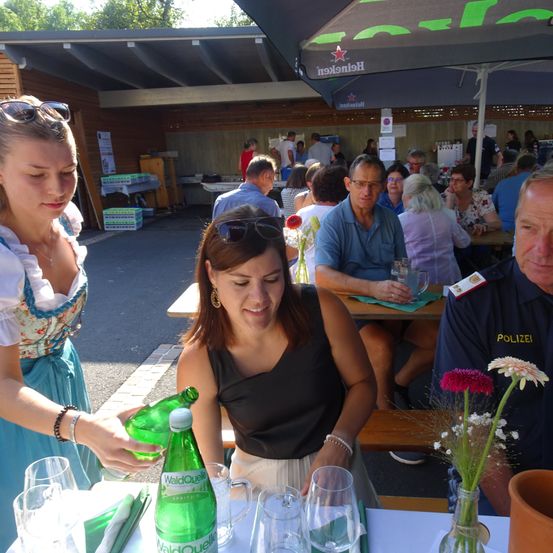 Eine Frau gießt Wasser aus einer grünen Flasche in ein Glas auf einem Tisch mit Blumen. Andere Menschen sitzen am Tisch und einige sitzen an anderen Tischen unter einem Schirm.