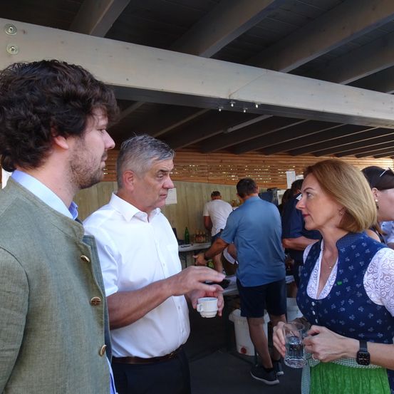 Ein Mann und eine Frau unterhalten sich mit einem älteren Mann bei einer Veranstaltung im Freien. Er hält eine Tasse, sie hält ein Glas Wasser. Andere Leute sind anwesend, darunter einer, der in der Nähe eines Tisches steht.