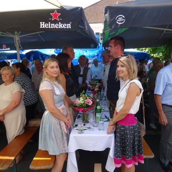 Eine Gruppe von Menschen steht an einem Tisch unter einem Heineken-Schirm. Zwei Frauen lächeln, während ein Mann zusieht. Auf dem Tisch stehen Blumen, Gläser und Flaschen.