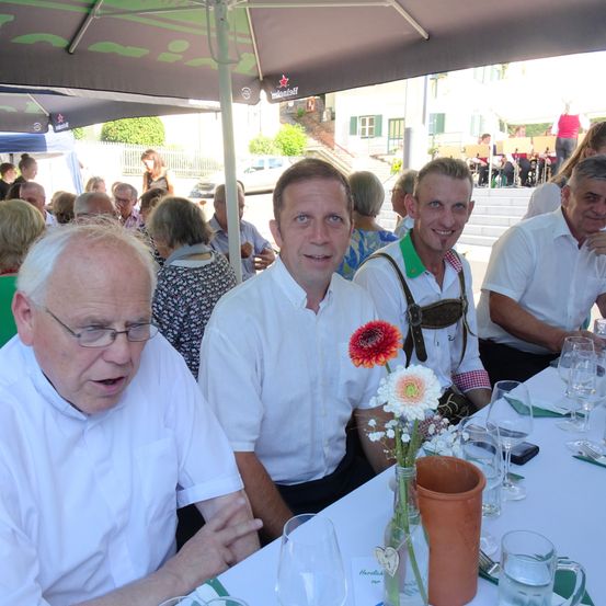 Drei Männer sitzen an einem Tisch unter einem Schirm, mit einem Blumenvase vor ihnen. Der Mann auf der rechten Seite trägt einen grünen Kragen und Hosenträger. Der Mann auf der linken Seite trägt eine Brille.
