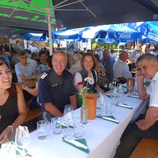 Eine Gruppe von Menschen, darunter ein Polizeibeamter, sitzt an einem langen Tisch in einem Biergarten unter Schirmen, lächelt und posiert für ein Foto.