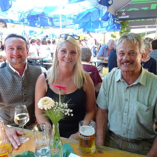 Drei Personen sitzen an einem Tisch unter blauen Sonnenschirmen. Ein Mann links und einer rechts lächeln. Die Frau in der Mitte hat Blumen in einer Vase. Hinter ihnen sitzen Leute an anderen Tischen.