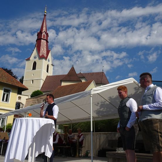 Ein Mann spricht in ein Mikrofon vor einer Kirche, während zwei andere Männer in der Nähe stehen. Dahinter spielt eine Band auf einer Bühne unter einem weißen Zelt.