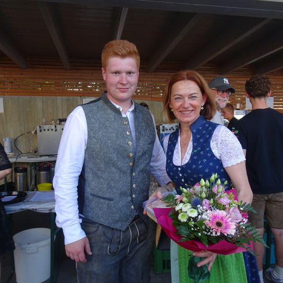 Ein Mann und eine Frau in traditioneller deutscher Kleidung lächeln für die Kamera und halten einen Blumenstrauß. Sie sind von einigen Leuten umgeben, von denen einer eine Kappe trägt. Hinter ihnen steht ein Tisch mit Behältern und einem Schreibtisch.