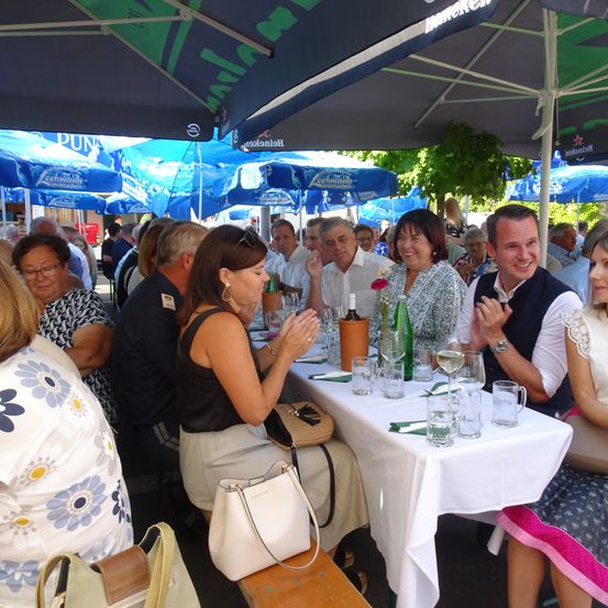 Eine Gruppe von Menschen sitzt um einen Tisch unter Schirmen, wahrscheinlich in einem Biergarten. Sie klatschen, lächeln und scheinen eine Mahlzeit zu genießen. Auf dem Tisch stehen Weingläser, eine Flasche und eine Blume.