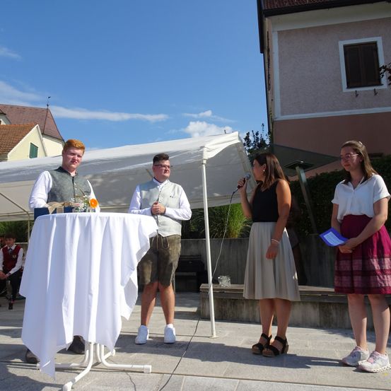 Drei Personen stehen vor einem Tisch unter einem Zelt. Ein Mann hält ein Mikrofon, während die anderen beiden neben ihm stehen. Ein Gebäude ist im Hintergrund.