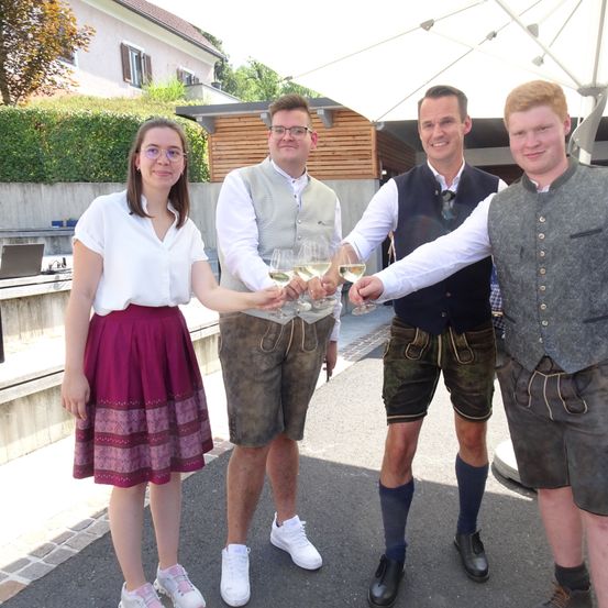 Vier Menschen stehen draußen. Zwei Männer tragen traditionelle deutsche Kleidung. Eine Frau trägt ein weißes Oberteil und eine lila Schürze. Sie alle halten Weingläser.