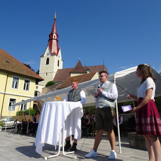 Zwei Männer stehen vor einem Tisch mit einem Mikrofon im Freien. Eine Frau steht neben ihnen. Hinter ihnen befindet sich ein Zelt und eine Kirche.
