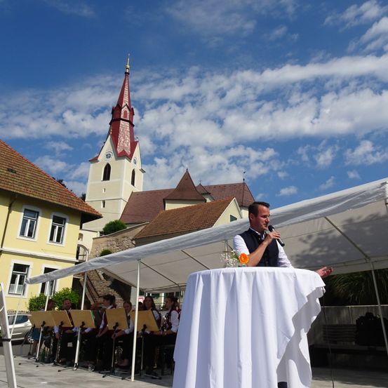 Ein Mann spricht in ein Mikrofon unter einem Zelt, mit einer Kirche im Hintergrund und einer Band, die darunter sitzt.