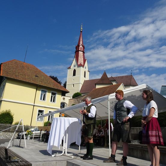 Drei Personen stehen vor einer Bühne, mit einem Zelt hinter ihnen und einer Kirche im Hintergrund. Der Mann auf der linken Seite hält ein Mikrofon.