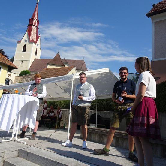 Eine Gruppe von Männern und Frauen steht auf einer Bühne, möglicherweise bei einem Festival. Ein Mann mit Mikrofon steht links. Ein Mann rechts hält eine Flasche. Hinter ihnen steht ein Zelt und eine Gruppe von Leuten sitzt auf der Bank.