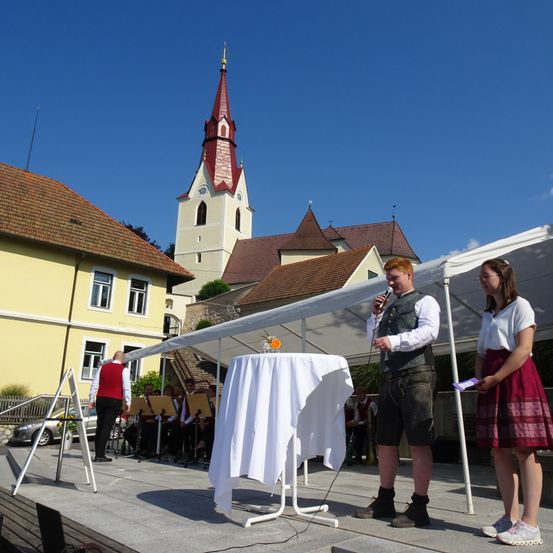 Ein Mann und eine Frau stehen auf einer Bühne vor einer Kirche. Im Hintergrund spielt eine Band.