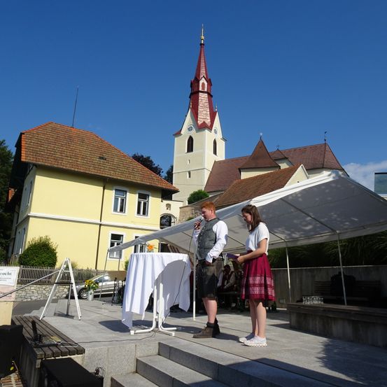 Ein Mann und eine Frau stehen auf einer Bühne vor einer Kirche, wahrscheinlich bei einer Aufführung. Die Bühne hat einen weißen Stoff, und der Mann hält ein Mikrofon. Hinter ihnen steht ein großes weißes Zelt. Die Kirche hat einen roten Turm und eine Uhr.