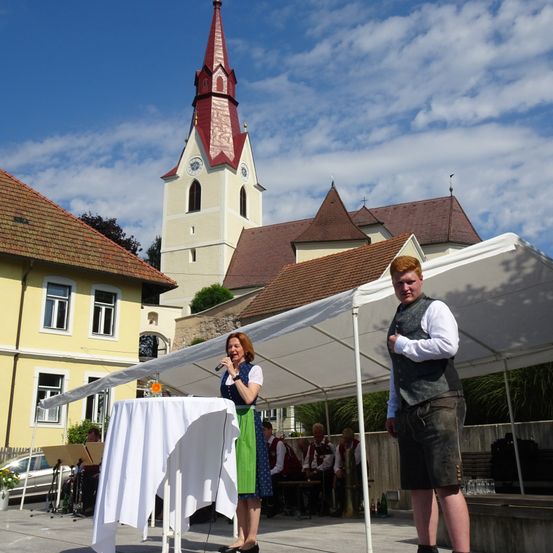 Eine Frau spricht in ein Mikrofon bei einer Veranstaltung im Freien vor einer Kirche. Ein Mann steht neben ihr. Dahinter befindet sich ein Vordach, ein Tisch mit weißer Tischdecke und eine Gruppe von Menschen, die auf einer Bank sitzen.
