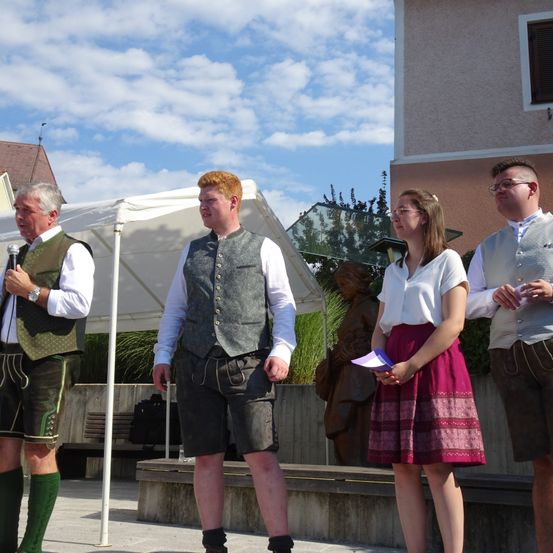 Vier Personen in traditioneller deutscher Kleidung stehen auf einer Bühne unter einem Zelt. Ein Mann hält ein Mikrofon, andere halten Papiere. Eine Statue und eine Bank sind im Hintergrund.