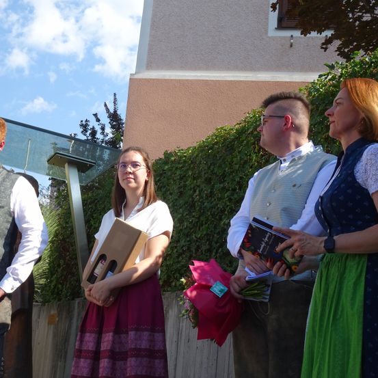 Drei Menschen stehen draußen vor einem Gebäude. Die Frau rechts hält einen Blumenstrauß und der Mann links eine Schachtel. Hinter ihnen steht eine Frau mit einem Buch.