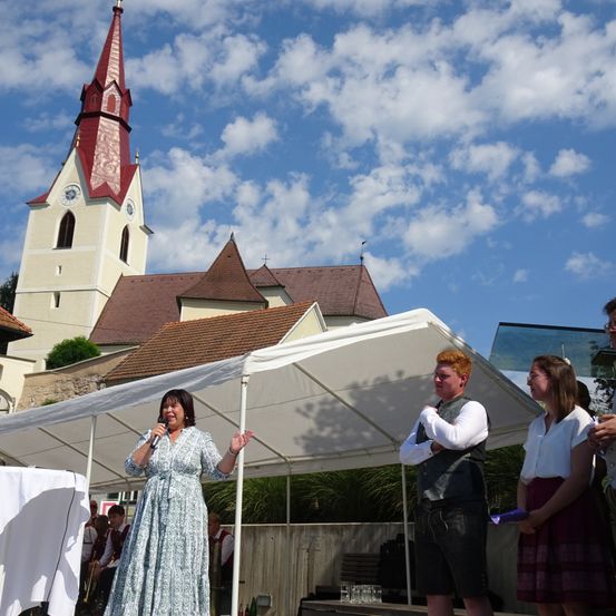 Eine Frau steht in einem Zelt, hält ein Mikrofon und spricht zur Menge. Hinter ihr stehen zwei Personen und schauen sich etwas an. Im Hintergrund befindet sich eine Kirche mit einem roten Dach und einer Uhr.