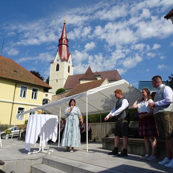 Eine Frau steht auf der Bühne unter einem Zelt und spricht in ein Mikrofon. Hinter ihr steht ein Tisch mit einer weißen Tischdecke. Vor ihr stehen zwei Männer und eine Frau auf den Stufen. Im Hintergrund befindet sich eine Kirche mit einem Turm und ein gelbes Gebäude.