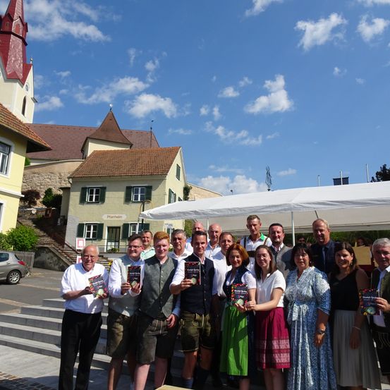 Eine Gruppe von Menschen posiert für ein Foto vor einem Gebäude mit einem Turm und einem großen weißen Zelt. Einige von ihnen halten Bücher.