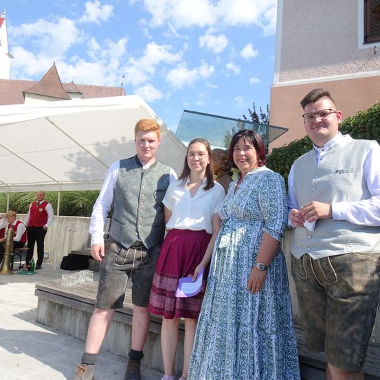 Vier Personen stehen in traditioneller deutscher Kleidung zusammen. Sie lächeln und scheinen für ein Foto zu posieren. Hinter ihnen ist ein Zelt und ein Gebäude zu sehen.