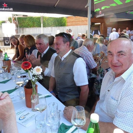 Eine Gruppe von Erwachsenen sitzt unter einem Regenschirm an einem Tisch und lächelt. Der Tisch ist mit Weingläsern, einer Blumenvase und Flaschen gedeckt. Der Mann im Vest schaut in die Kamera.