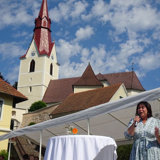 Eine Frau spricht in ein Mikrofon bei einer Outdoor-Veranstaltung vor einer Kirche unter blauem Himmel.